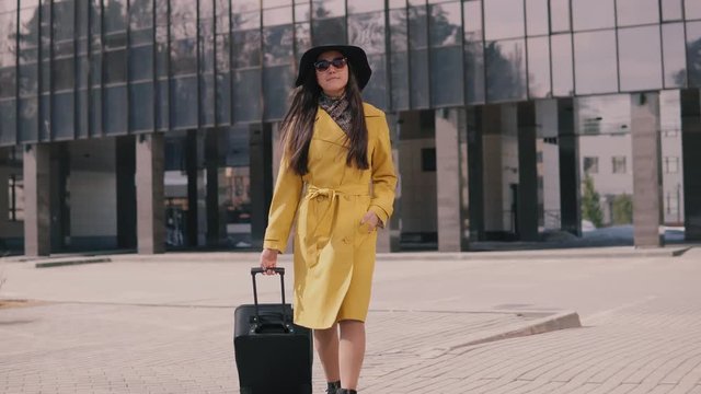 stylish young girl with a good mood in a yellow raincoat hat and glasses comes with luggage
