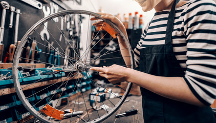 Cute Caucasian female worker holding and repairing bicycle wheel while standing in bicycle workshop.