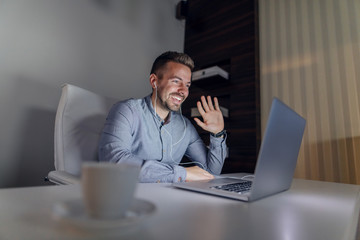 Cheerful handsome freelancer having video call over laptop. Man waving and having earphones in ears.