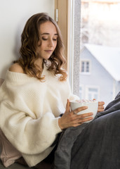 Girl sitting near the window, hold big white mag cacao with marshmallow. Home cozy lifestile photo