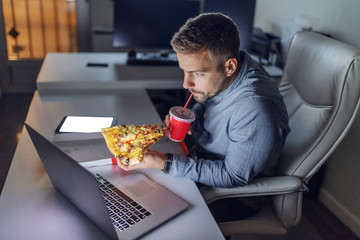 Young Caucasian architect drinking juice and eating pizza at office. On desk laptop. Late night...