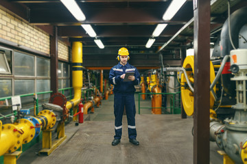 Full length of caucasian worker in protective suit, helmet and antifones on ears using tablet for...