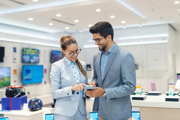 Smiling multicultural couple in formal wear trying out new smart phone while standing in tech store.