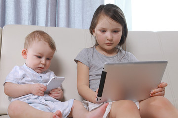 Children sit and play a game on the tablet and phone, boy and girl enthusiastically looking at the screen