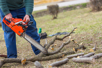 Old man holding orange chainsaw with his bare hands and cutting a branch placed on the ground. Orange chainsaw in action.
