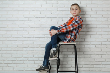 Cute stylish boy near white brick wall . fashionable boy sitting on chair near brick wall. Portrait of a funny boy in jeans and a shirt in a cell.