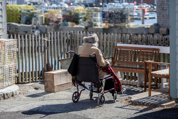 Obraz premium An elderly woman sits in her wheelchair warmly dressed outside in a New England sea side setting.