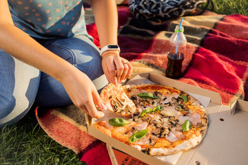 Happy young woman holding hot pizza in box, seat outdoor in park