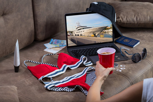 Preparing For Vacation. Woman's Hand With A Red Plastic Cup Of Soda On The Background Of An Open Modern Laptop Marks The Beginning Of The Tour. Beach Holiday.