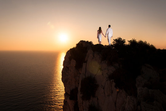 Couple in love holding by hands, standing on rocky cliff and looking on breathtaking landscape with sunset over sea.