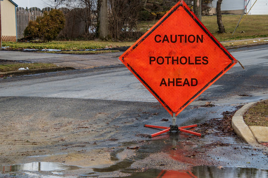 Orange Triangular Road Sign On A Small Suburban Street That Says Caution Potholes Ahead