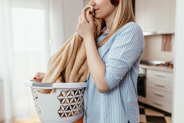 Beautiful young smiling woman is smelling clean towels while doing laundry at home.