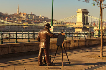 Budapest, Hungary-January 08, 2017:Copper statue of the painter who paints the landscape of the city of Budapest. Chain Bridge across River Danube and Medieval Fisherman's Bastion in the background