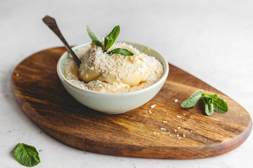 Homemade vegan ice cream in a bowl is made from frozen mango and coconut milk served by fresh mint and flakes on a wooden textured board.