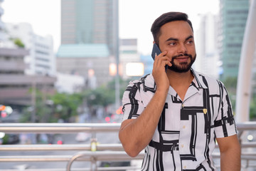 Happy young bearded Indian man talking on the phone at the footbridge