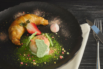 Slice of apple strudel with a scoop of vanilla ice cream on a black plate, on a wooden black table, decorated with meringue and strawberries. close-up. horizontal view of baked dessert.
