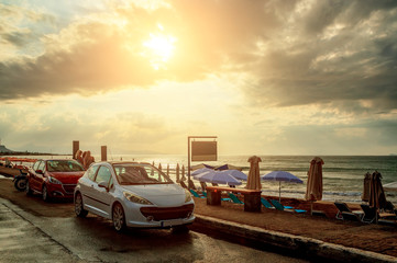 beige tinted cars on the waterfront by the sea with the beach and umbrellas in the background of the sunset and clouds. Crete, Greece