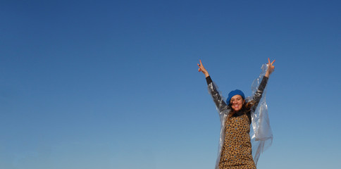 girl with her hands up looks at the camera and smiles. Dressed in a dress, a transparent raincoat and a blue hat. Against the blue sky. Day. Spring, summer or autumn. Horizontally