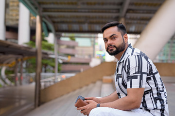 Obraz premium Young bearded Indian man thinking with phone on staircase at the city