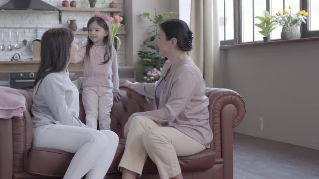 Grandmother, mother and little daughter spend time together sitting on the sofa in modern apartment. The girl standing on the cout, mom and granny looking at her smiling. Concept of generation.
