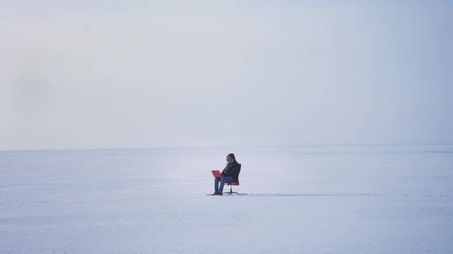 Wide shot of alone businessman in warm coat and hood sitting in snow desert and working with laptop attentively.
