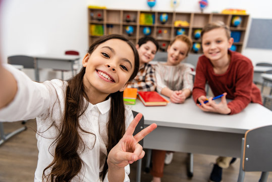 Cheerful schoolgirl taking selfie with friends and showing peace sign in classroom - Powered by Adobe
