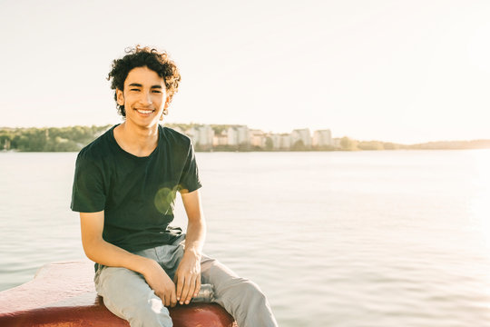 Portrait of smiling teenage boy sitting on bollard by river