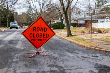 Orange triangular road sign on a small suburban street that says Road Closed Potholes