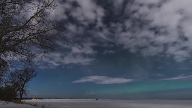 Time-lapse of moonlit clouds blowing over a snow covered lake with aurora dancing in a starry sky. 
