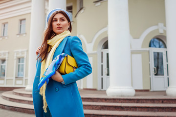 Young woman in trendy blue coat walking in city holding stylish handbag. Spring female clothes and accessories. Fashion