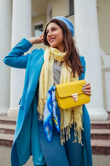 Young woman in trendy blue coat walking in city holding stylish handbag. Spring female clothes and accessories. Fashion