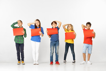 Obraz premium Group of saddened and distressed children with red empty banners isolated on white studio background. Education and advertising concept. Protest and children's rights concepts.
