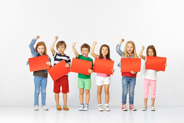 Obraz premium Group of unhappy angry children with red empty banners isolated in white studio background. Education and advertising concept. Protest and children's rights concepts.