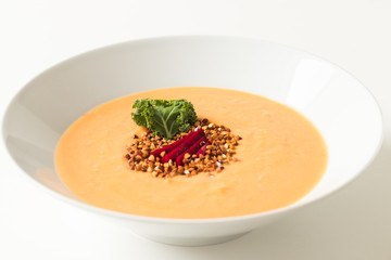 Bowl of pumpkin cream soup decorated with crunchy buckwheat, beetroot and kale salad on white background.