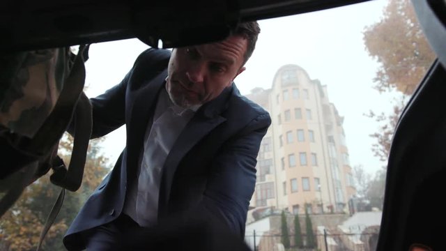 Serious businessman puts his stuff in the boot of the car. Volunteering, military backpack, going home. Tired, exhausted, depressed. Hard times, war conflict, safety, helping the others