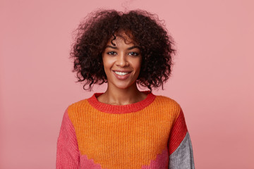 Indoor portrait of positive optimistic charming african american girl with afro hairstyle looks with pleasure, with lively smile, wearing colorful longsleeve, isolated on pink background - obrazy, fototapety, plakaty