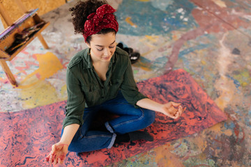 Young beautiful woman with closing eyes sitting on yoga mat in lotus pose and meditating dreamily spending time in big workshop with paint splashes around