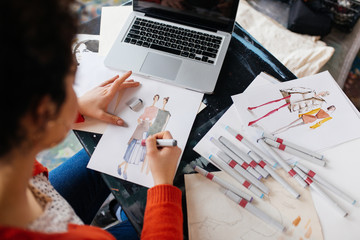 Top view of young woman sitting at the table with laptop dreamily drawing fashion illustrations while spending time in modern workshop
