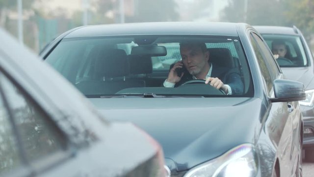 Angry nervous bearded man in luxury suit stands in traffic jam, picks up his phone, answers call, explaining situation, actively gesticulating. Being in a hurry, being late, stressful situation