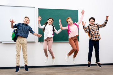 Four pupils with backpacks jumping in front of blackboard