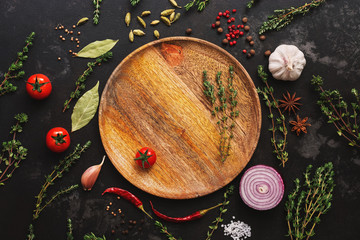 Obraz na płótnie Canvas Different seasoning for cooking on a dark background. Empty wooden plate, spices, herbs, vegetables. Top view, flat lay.