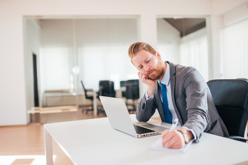 Fototapeta premium Tired redhead businessman at office desk, taking notes.