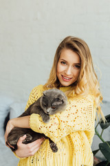 beautiful smiling girl in knitted sweater looking at camera and hugging cute scottish fold cat