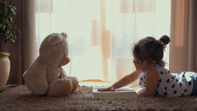 Little girl lying on the floor reading a book to a toy teddy bear