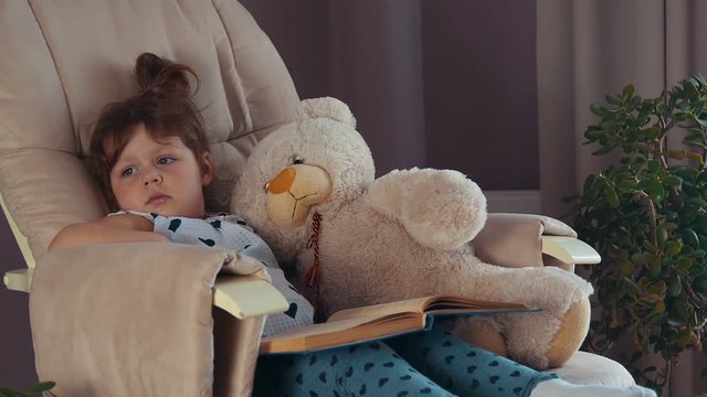 Little girl waking up on the chair with big book hugging toy teddy bear
