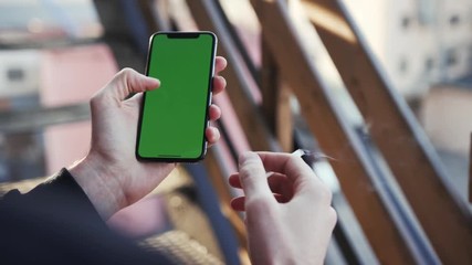 Smartphone with green display in vertical position being used by actively gesticulating male hands in the same time smoking cigarette. Technology, android basis, flagman, scrolling pages, tapping