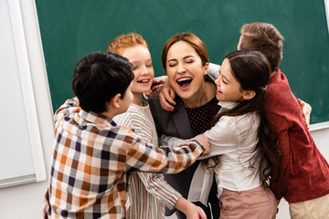 Happy pupils embracing teacher in front of blackboard in classroom