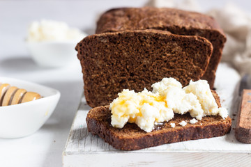 Slice of dark rye bread with fresh farm cheese and honey on a white wooden cutting board.