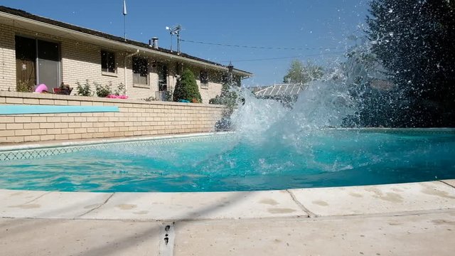 Young fit Caucasian man runs off diving board and jumps into a swimming pool in a backyard. Man in swimming trunks jumps into an outdoor swimming pool. Jumping into backyard pool making a splash.