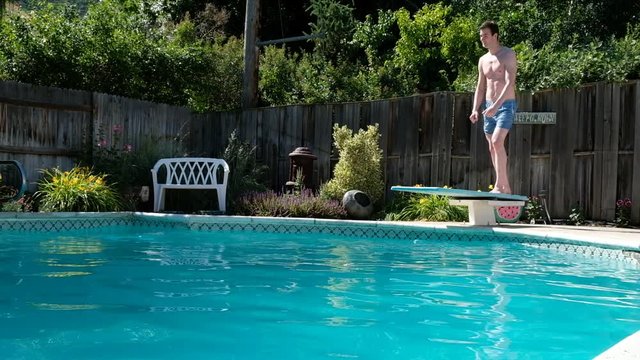 Young fit Caucasian man runs off diving board and jumps into a swimming pool in a backyard. Man in swimming trunks jumps into an outdoor swimming pool. Jumping into a private backyard pool.